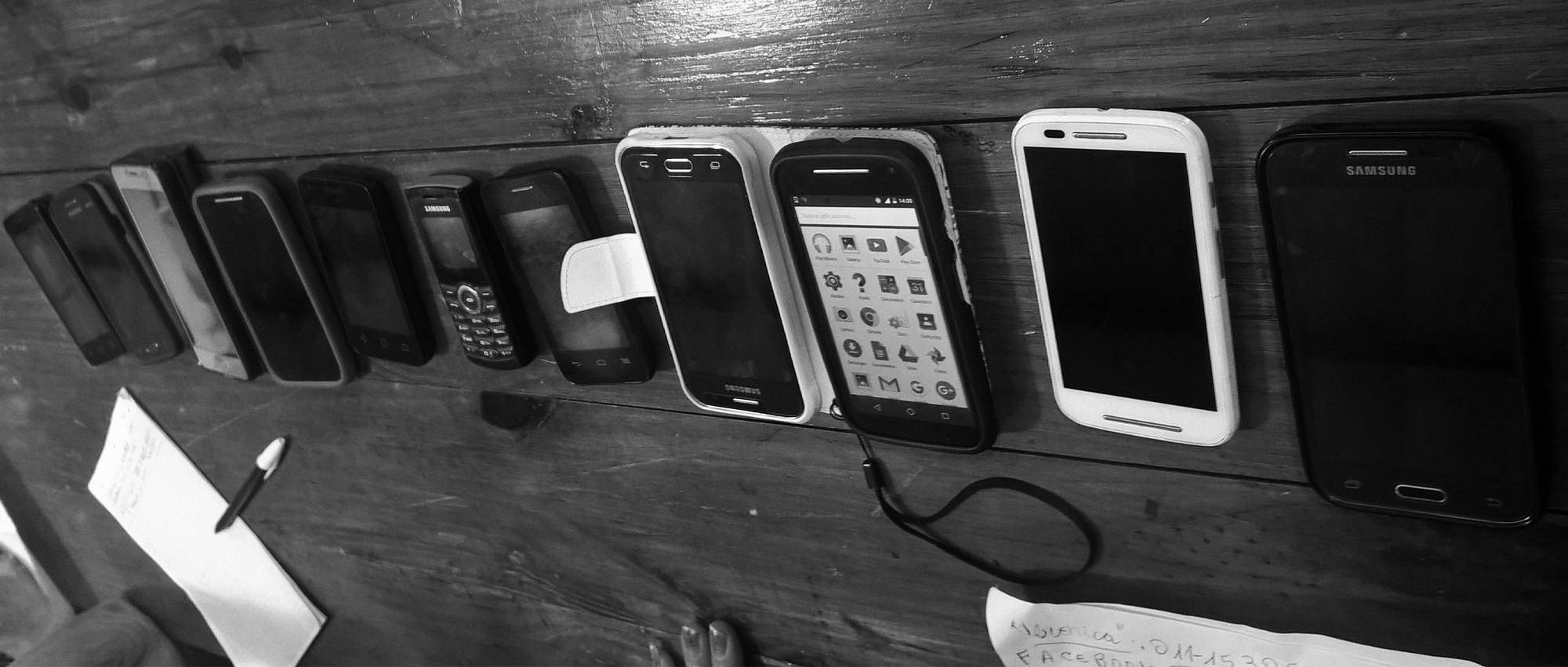 Old smartphones of diverse models on a table next to pens and notes.